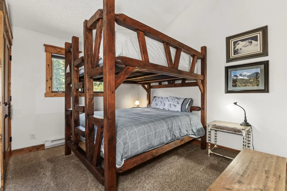 This image features a welcoming bedroom with a Queen-over-Queen bunk bed setup, complemented by a nightstand and a cozy desk lamp. Framed pictures add a personal touch, and the inviting mood is perfect for both adults and children. The arrangement offers ample sleeping space and fosters a sense of fun and comfort.