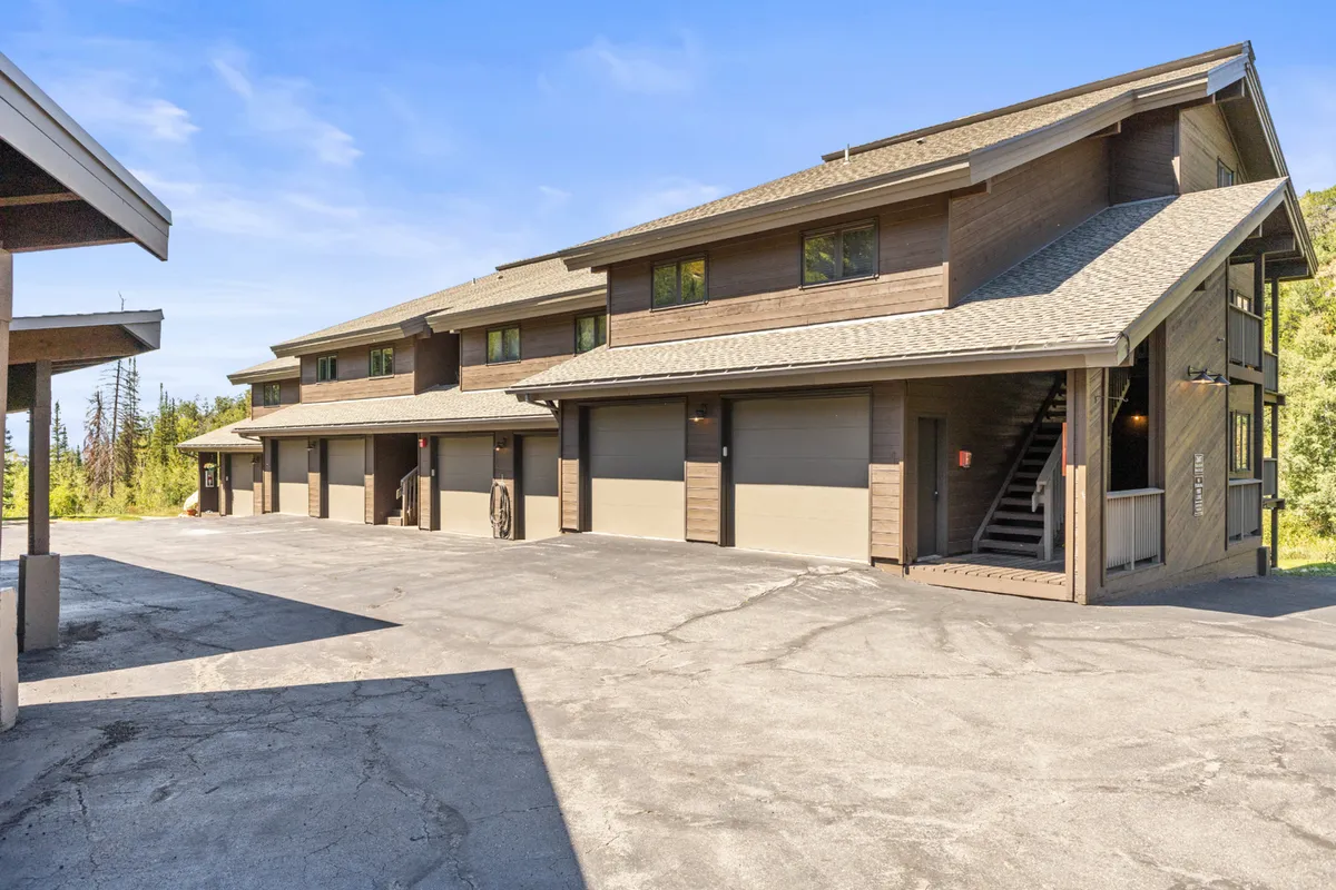 A wide exterior shot showcasing the multi-unit building with garage doors and a stairway entrance, surrounded by serene landscaping. The setting suggests community, accessibility to outdoor activities, and an inviting environment for residents and guests alike.