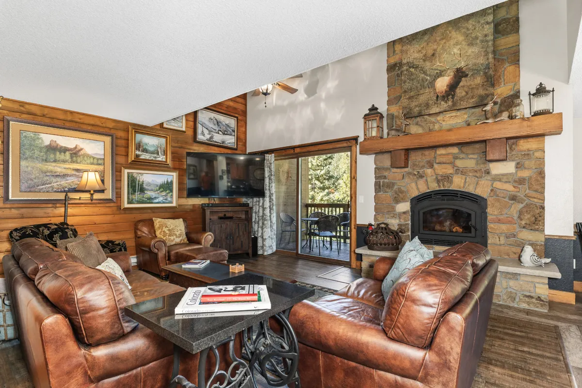 The image showcases a beautifully renovated condo living room with a striking stone fireplace as the centerpiece, surrounded by plush leather sofas and tasteful wooden wall art. Sunlight pours in through a large window, offering a glimpse of the natural beauty of Steamboat Springs right outside.