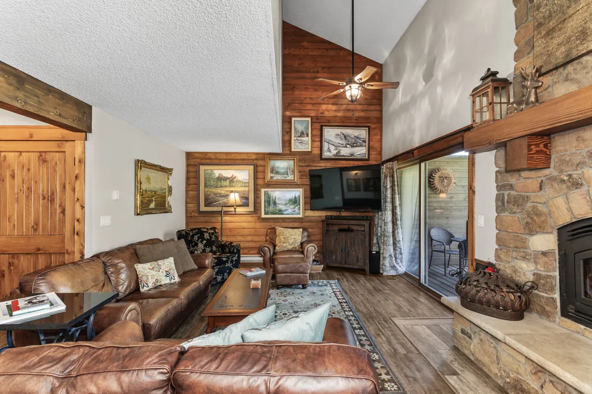A warm and inviting living room featuring plush leather sofas arranged around a striking stone fireplace. Wood paneling and framed artwork add rustic charm, while a large window lets in ample natural light and offers a hint of the outdoors.