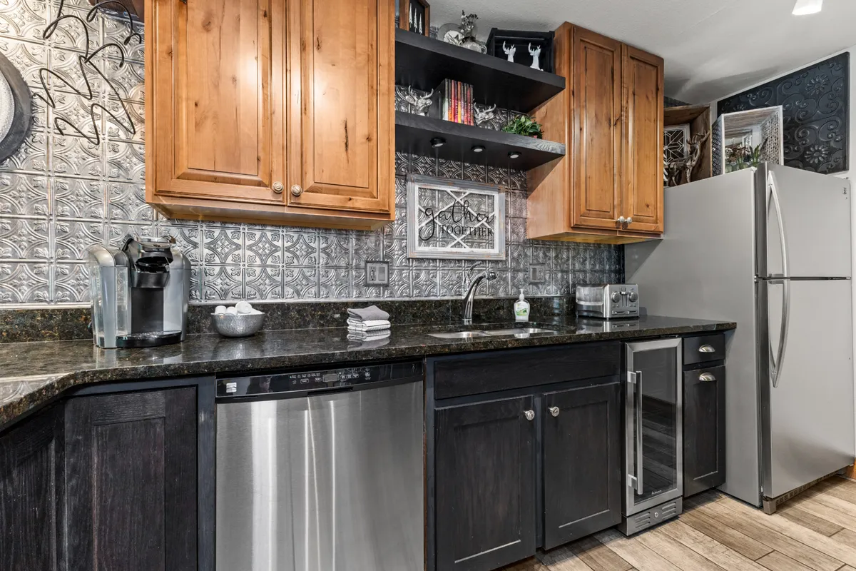 A spacious kitchen area showcasing a granite countertop, wood cabinets, open decorative shelves, and stainless steel appliances. The open layout encourages socializing and group meal prep.